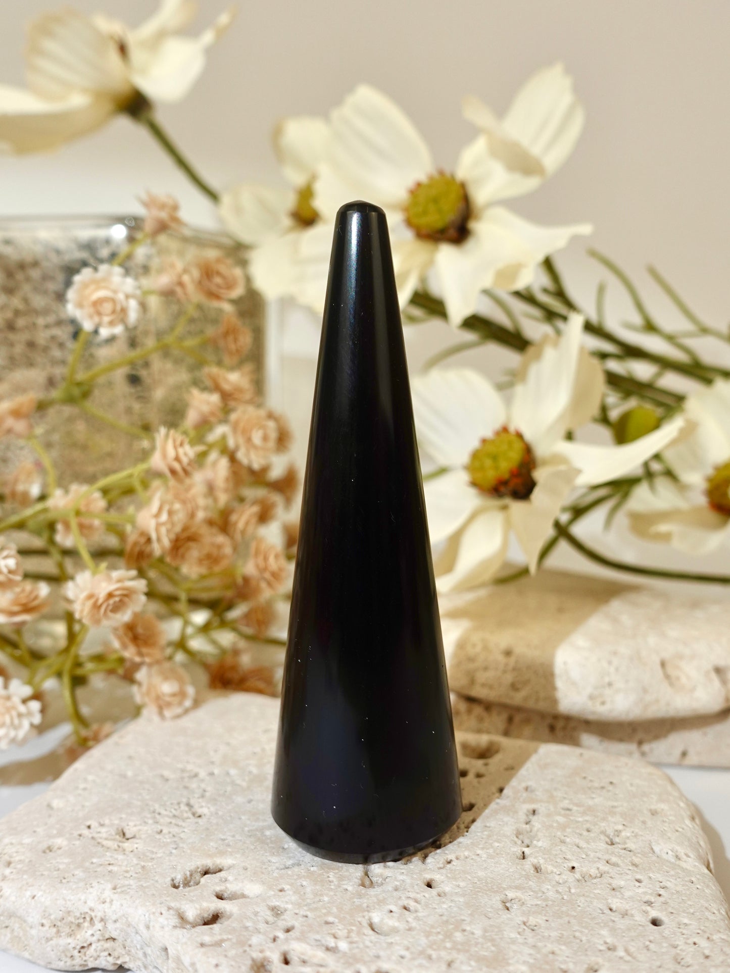 A glossy black cone-shaped crystal or mineral specimen displayed upright on a light beige stone slab, with dried white flowers and neutral stones in the softly lit background.