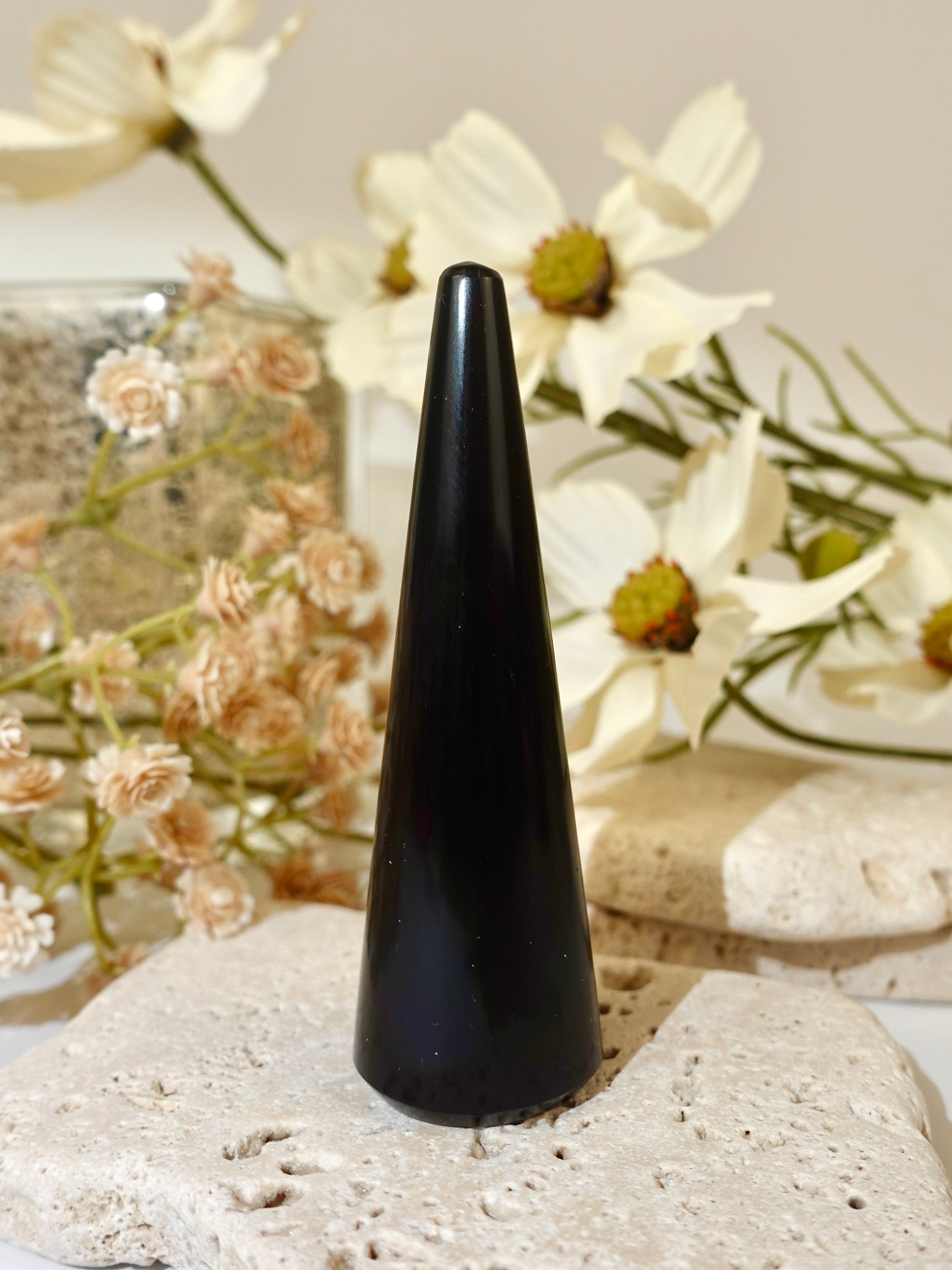 A glossy black cone-shaped crystal or mineral specimen displayed upright on a light beige stone slab, with dried white flowers and neutral stones in the softly lit background.