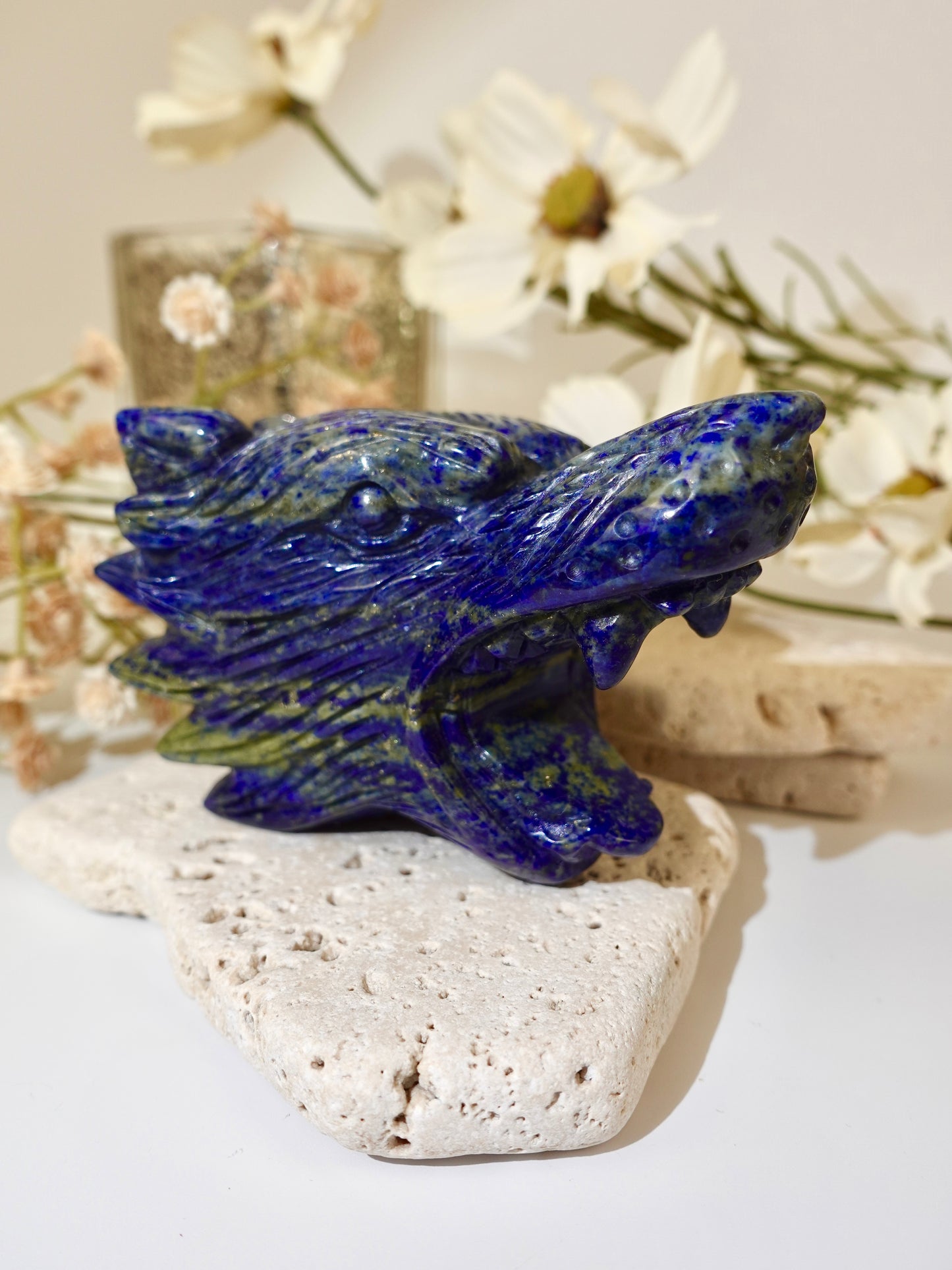 A carved blue lapis lazuli wolf figurine displayed on a light stone slab with dried white flowers in the background.