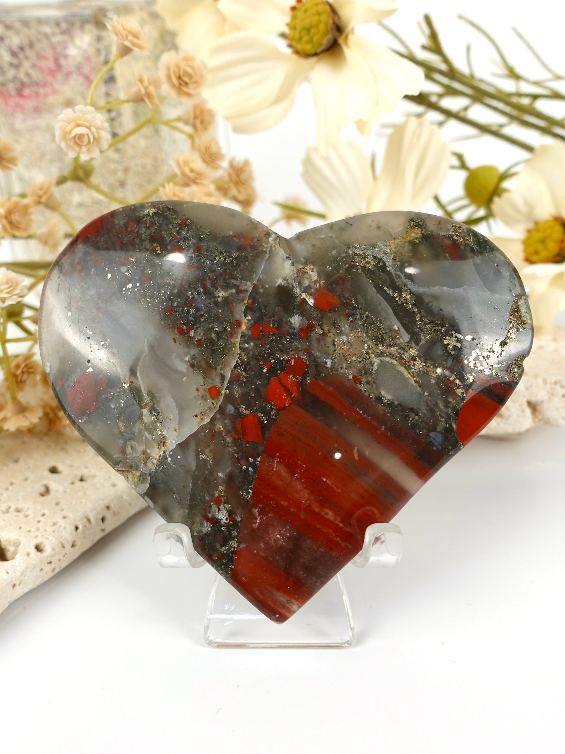 A heart-shaped polished stone display piece on a clear acrylic stand, featuring natural banding and speckling in gray, black, white, and red, set against a soft-focus background of dried flowers.