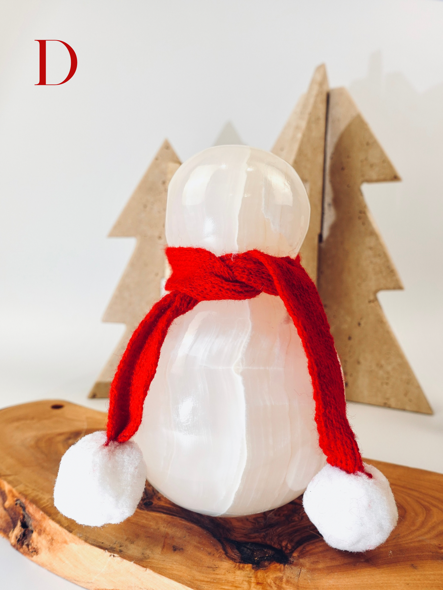 Snowman figurine with a red scarf on a wooden surface with Christmas tree decorations in the background.