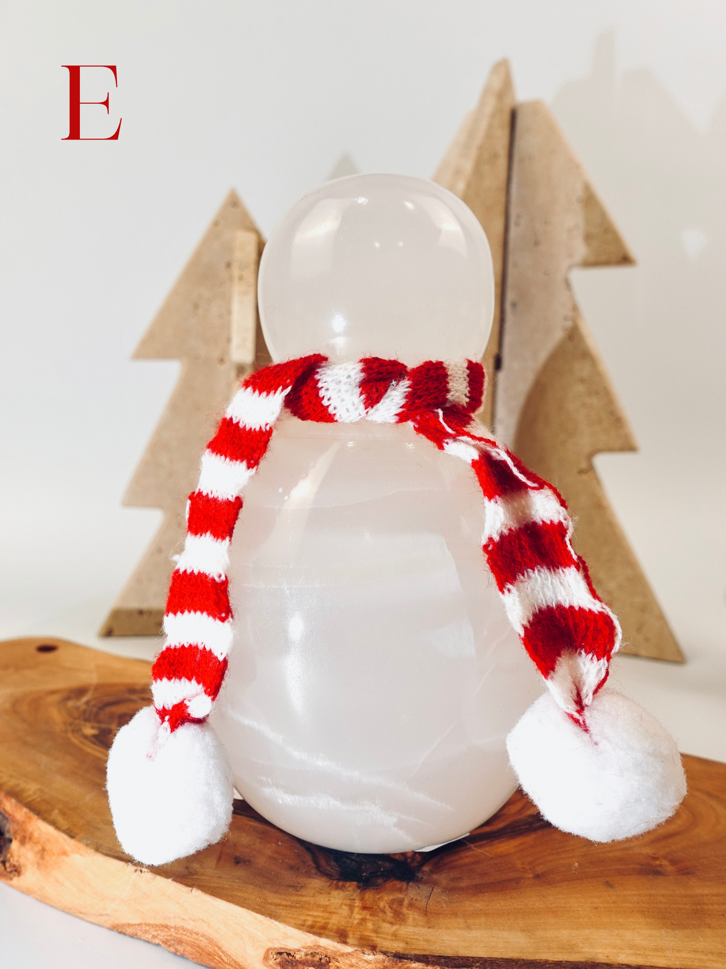 Snowman figurine with a red and white striped scarf on a wooden surface with Christmas tree decorations in the background.