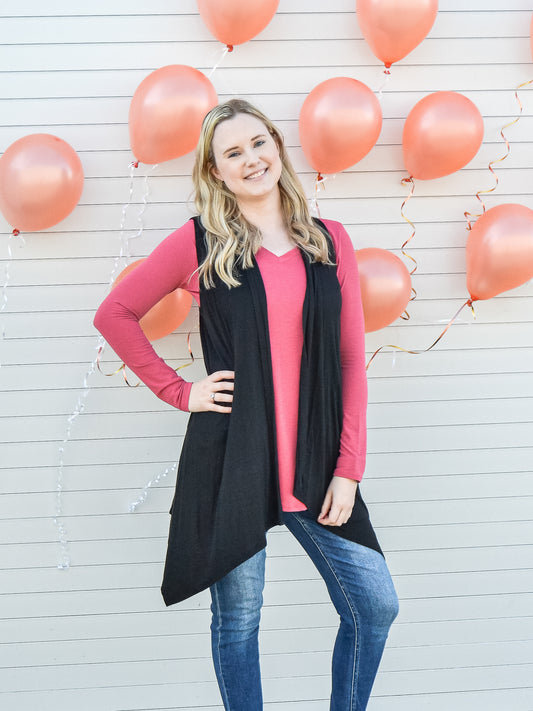Black open front sleeveless cardigan styled with pink long sleeve and jeans.