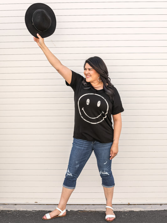 Black, short sleeve graphic tee with a smily face. Styled with capris and a hat.